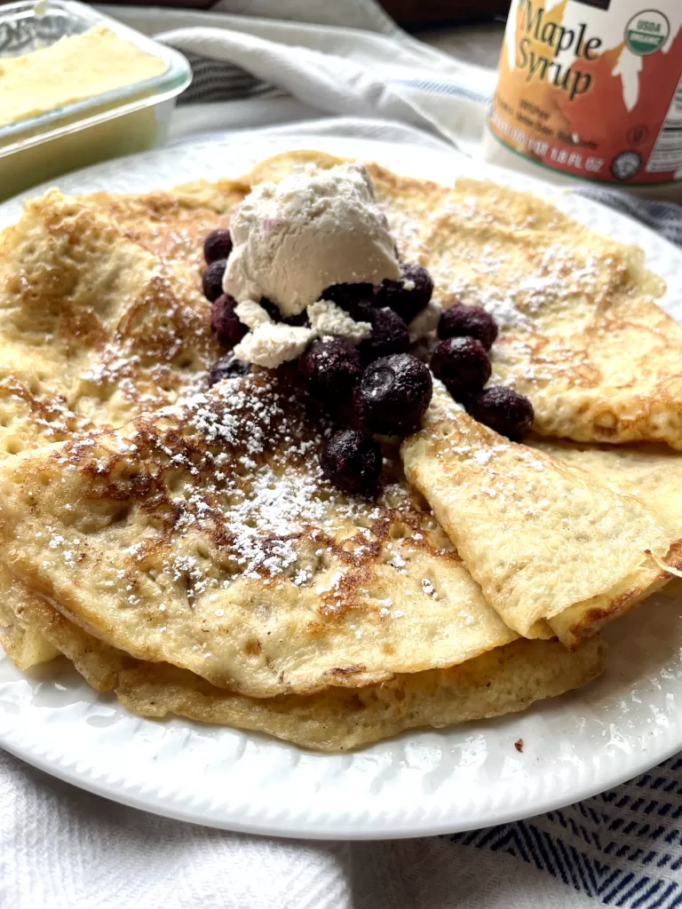 cooked sourdough crepes on a white plate topped with whipped cream, blueberries, and powdered sugar