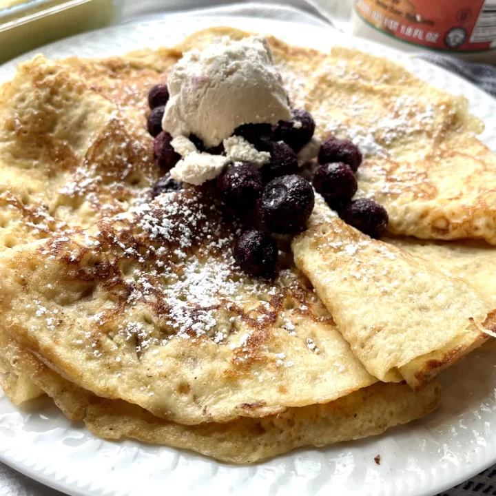 cooked sourdough crepes on a white plate topped with whipped cream, blueberries, and powdered sugar