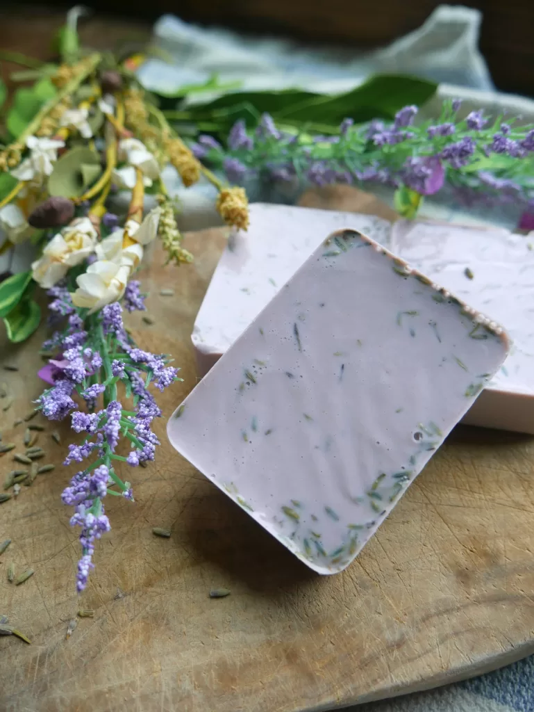 homemade lavender soap bar propped up on other lavender soap bars next to purple flowers