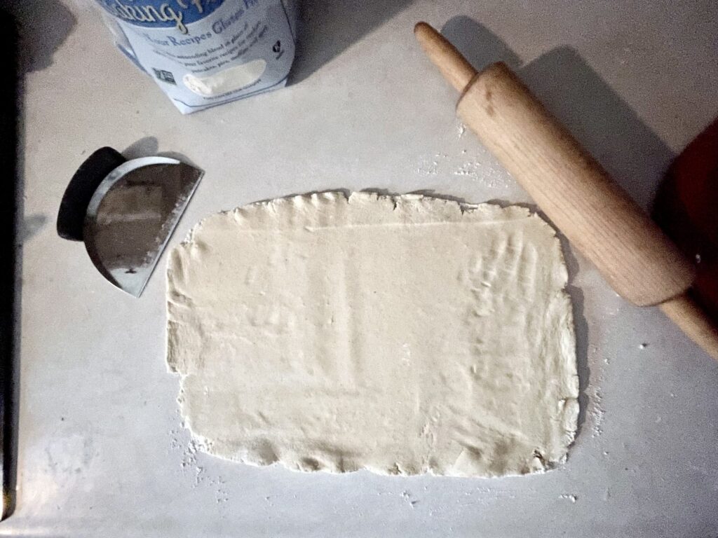 rectangular gluten free cinnamon roll dough rolled out on the counter next to a rolling pin and bench scraper