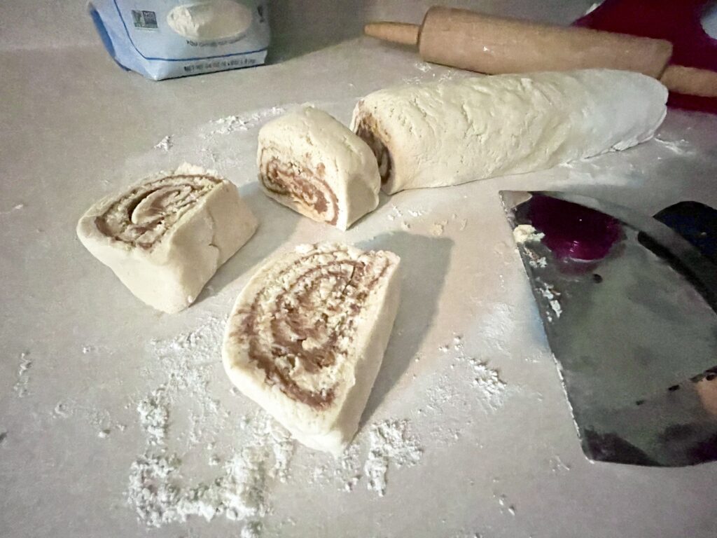 3 unbaked gluten-free cinnamon rolls sitting next to a bench scraper and rolling pin on white counter