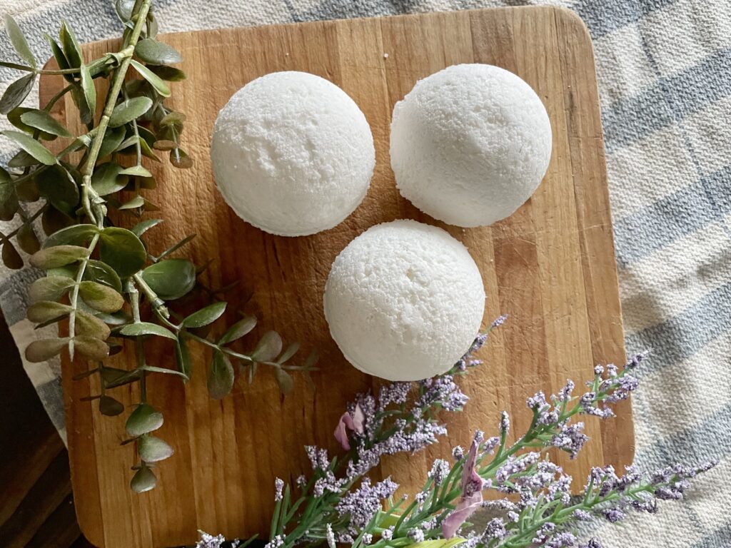 3 bath bombs made without cornstarch sitting on a wooden cutting board with purple flowers and green leaves around them