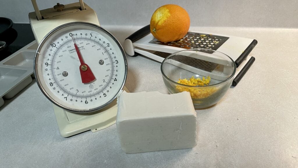 melt and pour soap base, an orange on a grater, and a scale