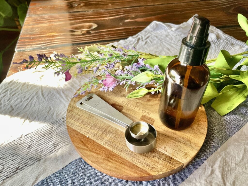 DIY body oil in brown glass bottle next to measuring spoons on a wooden trivet with florals