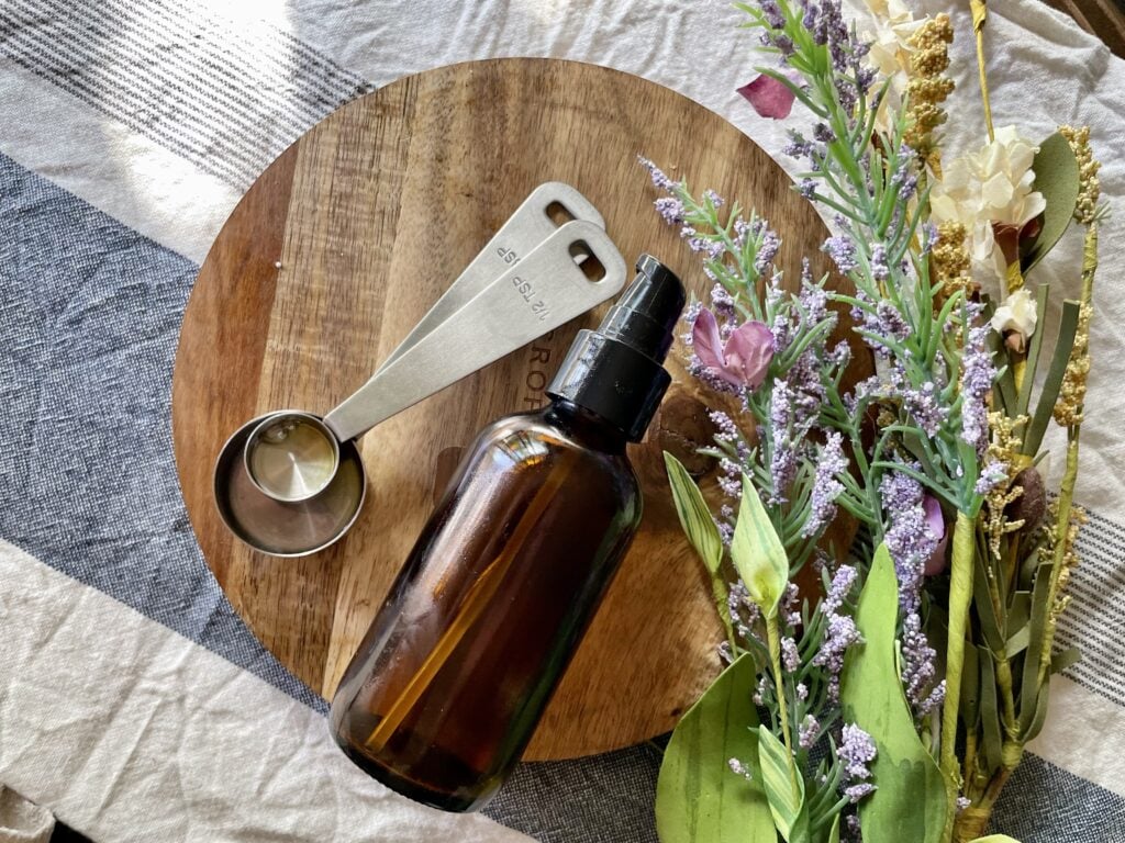 homemade body oil in brown glass bottle next to metal measuring spoons and flowers