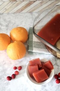 homemade jello sliced in a small white bowl with large glass pan of healthy jello in background next to oranges and cranberries