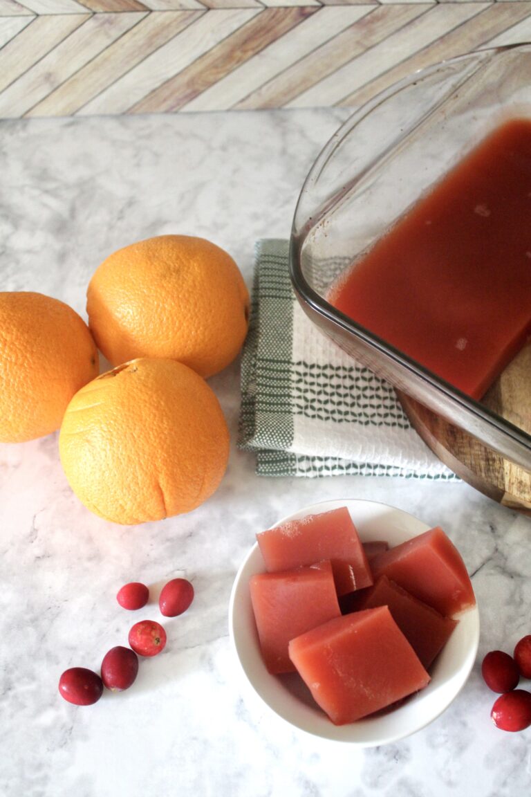 homemade jello sliced in a small white bowl with large glass pan of healthy jello in background next to oranges and cranberries