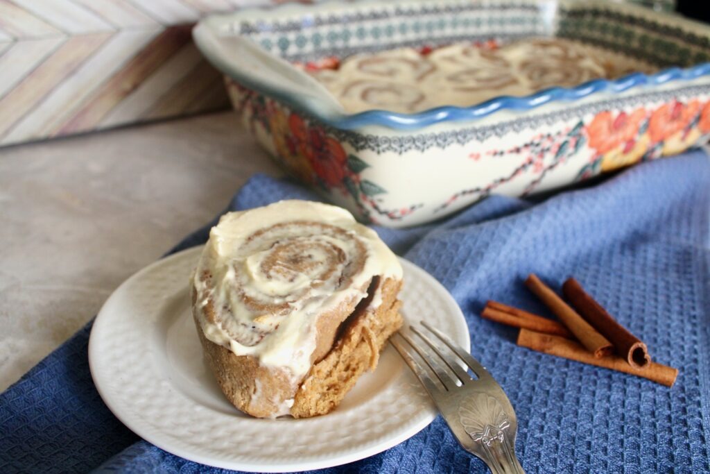 one freshly milled flour sourdough discard cinnamon roll on white plate in front of pan of cinnamon rolls